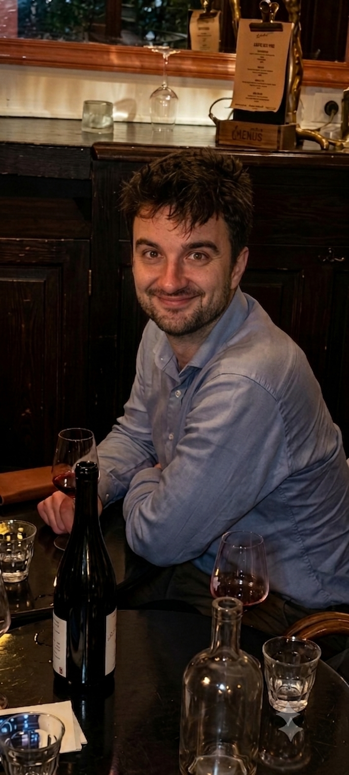 Arthur attablé dans un bistrot, chemise bleue, sourire, un verre et une bouteille de vin rouge devant lui